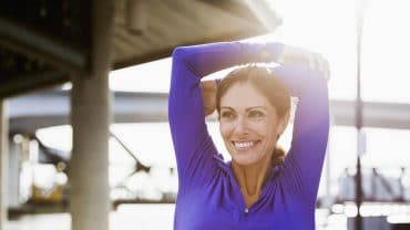 Mulher madura, fazendo uma pausa na corrida para esticar os braços. Ela está ao ar livre em uma passarela de pedestres à beira-mar. É um dia claro e ensolarado. Ela está sorrindo, desviando o olhar da câmera.