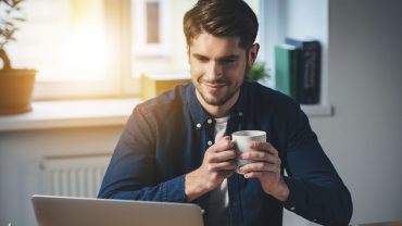 homem segura uma xícara de café enquanto olha para a tela do computador. Ele está concentrado e sorri timidamente