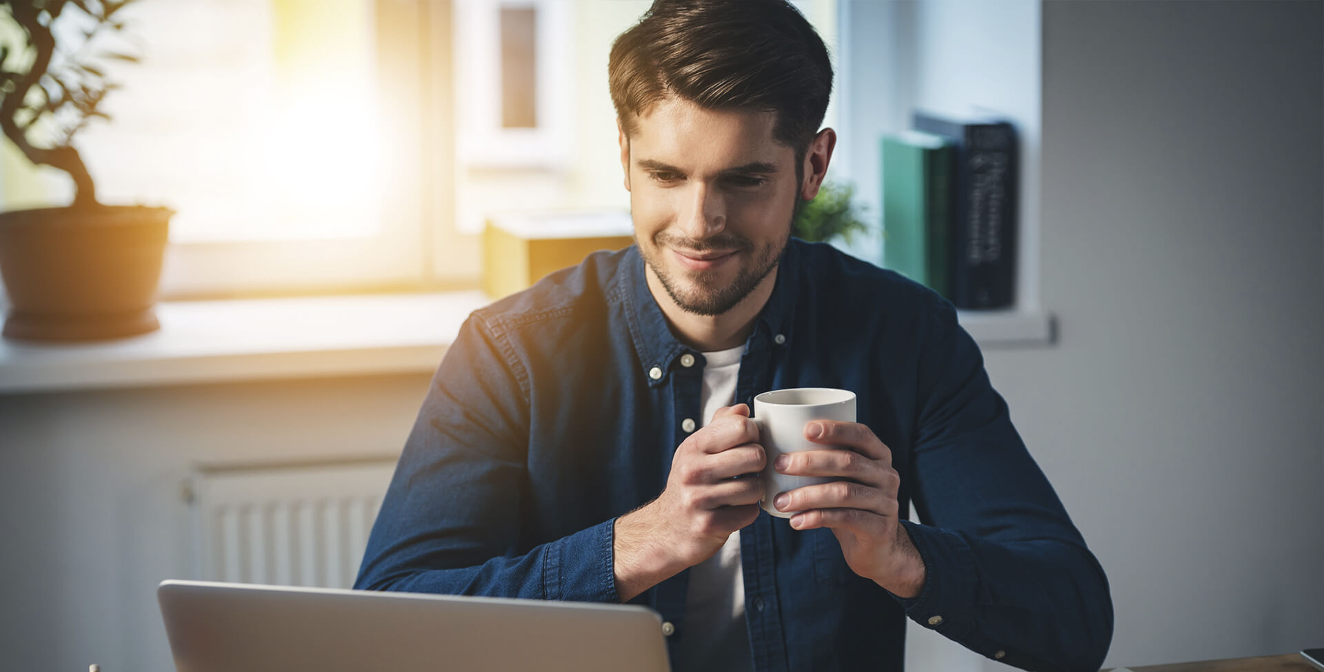 homem segura uma xícara de café enquanto olha para a tela do computador. Ele está concentrado e sorri timidamente