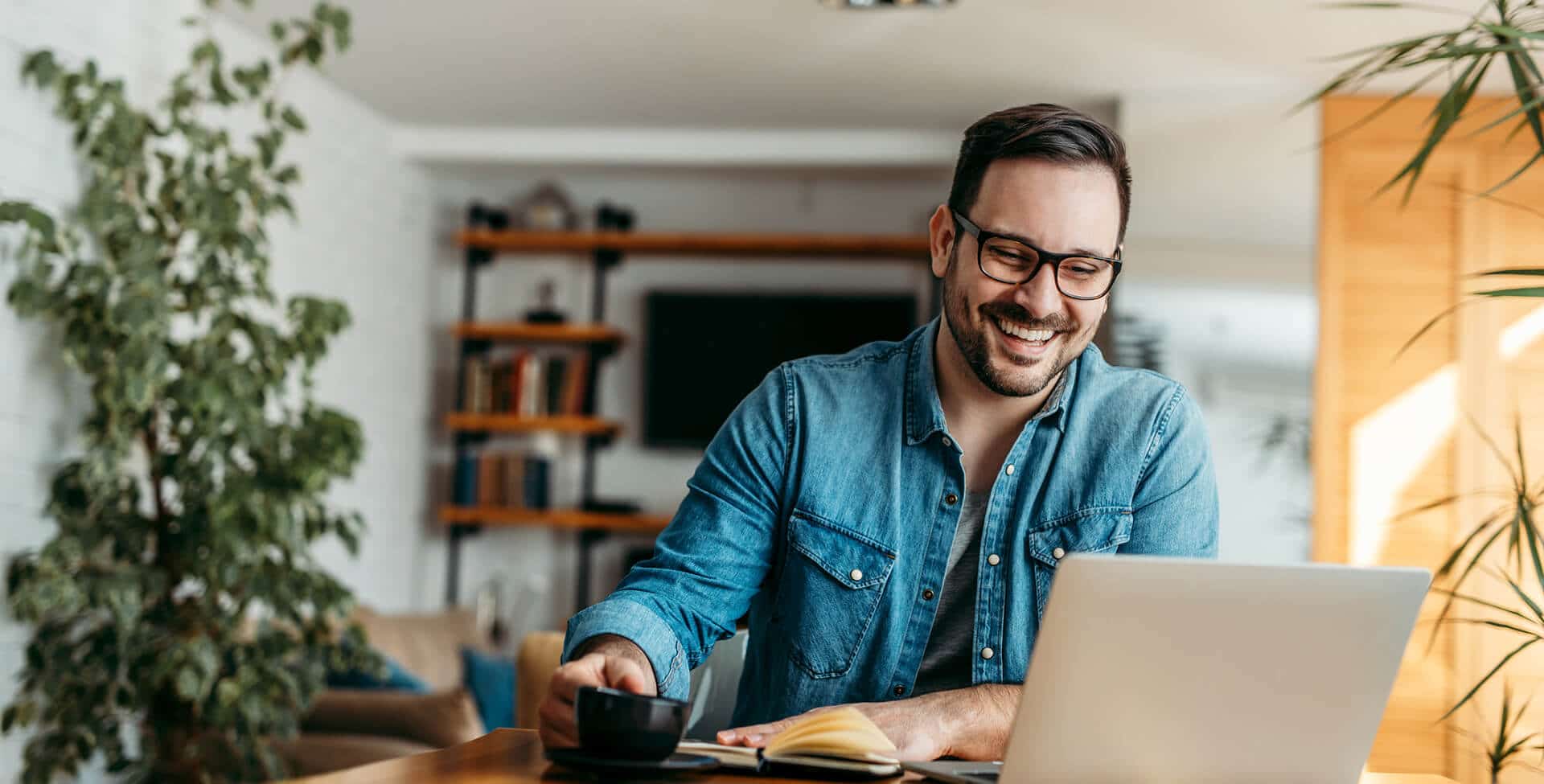 homem sorri em home office, olhando para laptop e bebendo xícara de café.