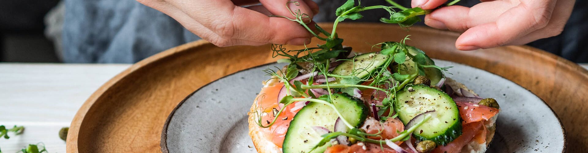 a imagem mostra as mãos de uma mulher preparando uma torrada com alimentos saudáveis, como salmão, pepino. Ela coloca brotos em cima da torrada, que está em um pratode cerâmica e um souple de madeira, sob uma mesa