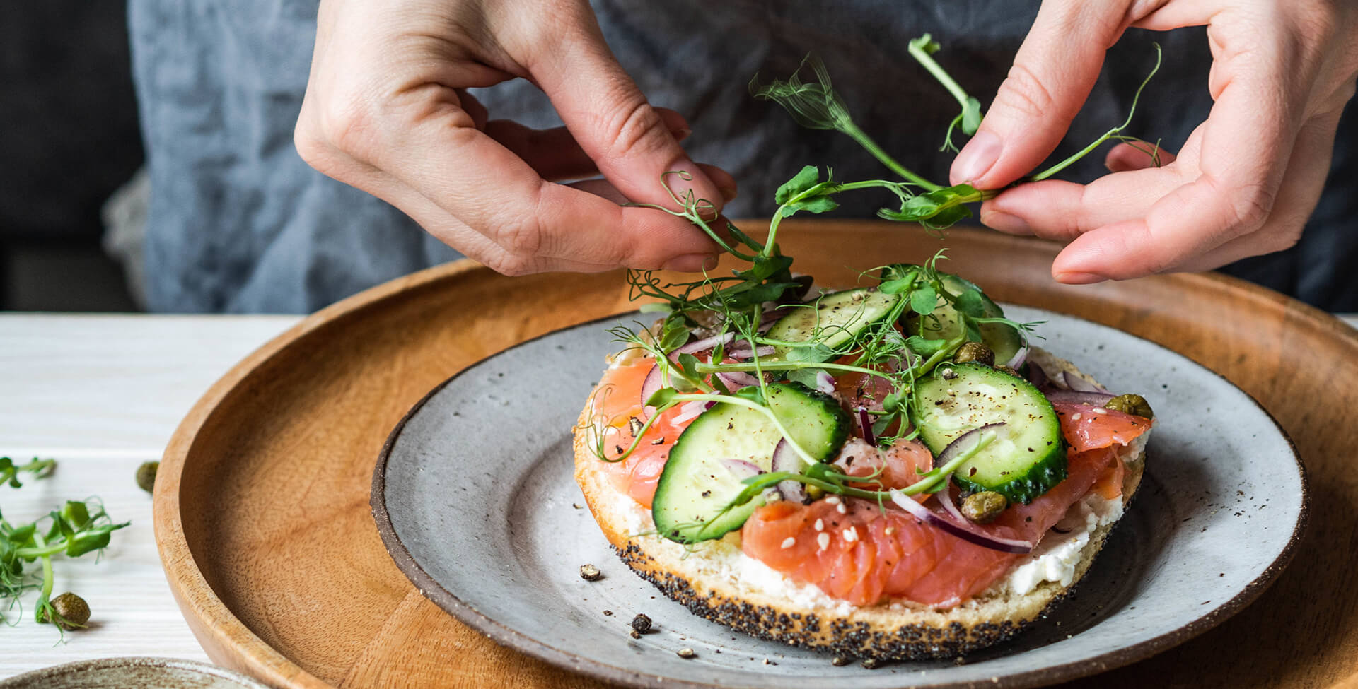 a imagem mostra as mãos de uma mulher preparando uma torrada com alimentos saudáveis, como salmão, pepino. Ela coloca brotos em cima da torrada, que está em um pratode cerâmica e um souple de madeira, sob uma mesa